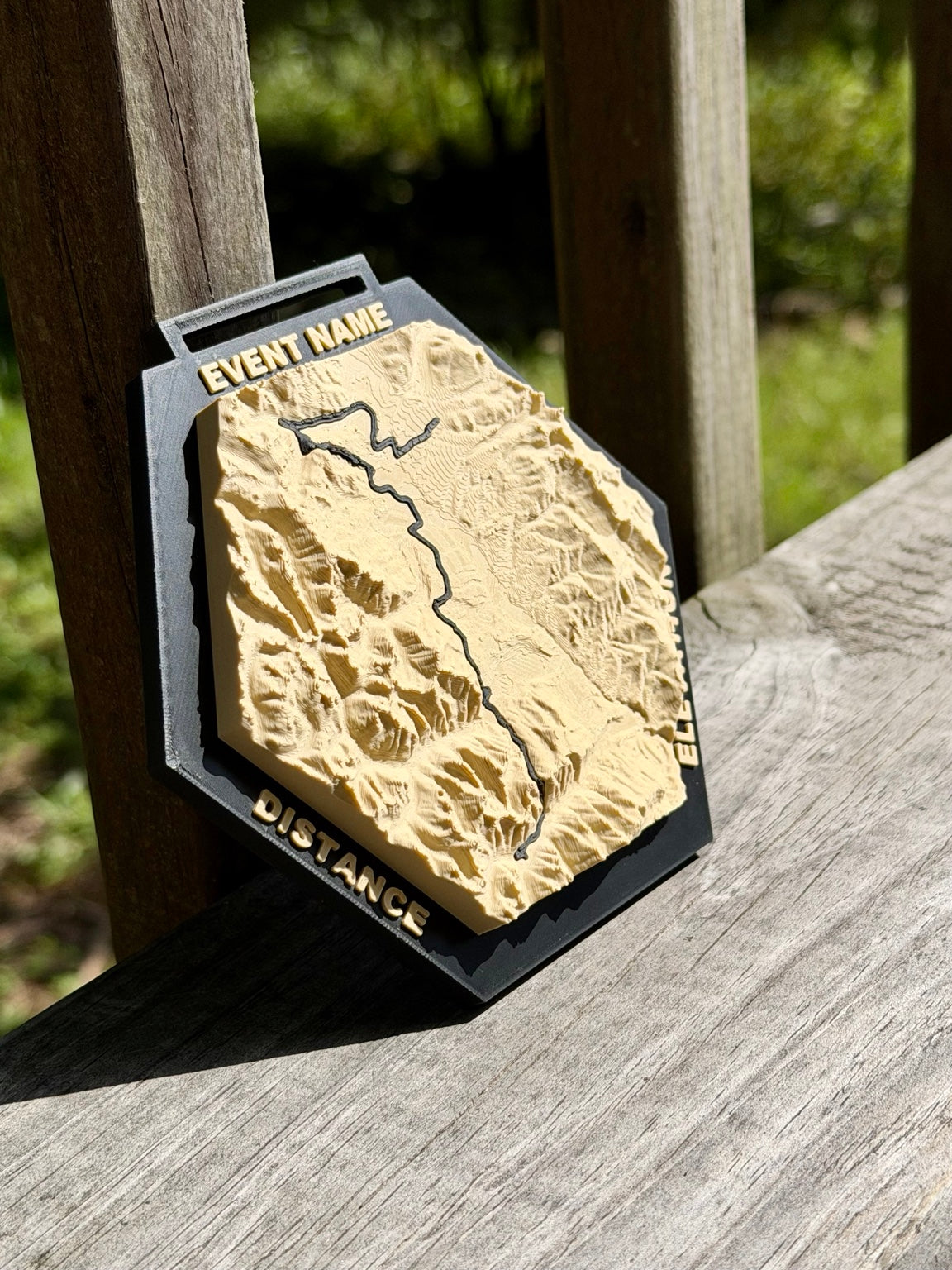 Angled view of a Race Topos 3D topographic medal showing raised terrain and lettering on a black hexagonal frame.
